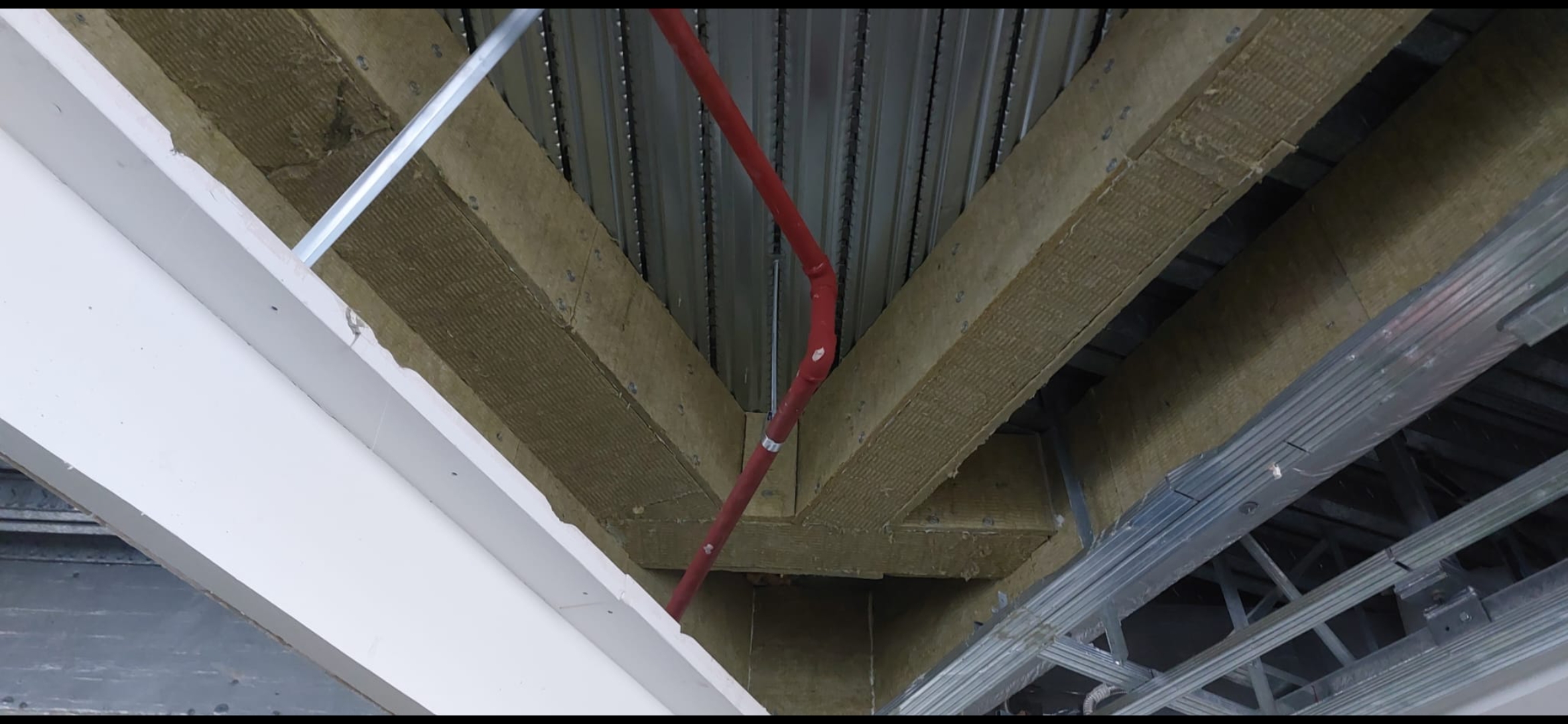 Red fire-stopping pipe penetration through mineral wool insulation at a steel deck ceiling junction.