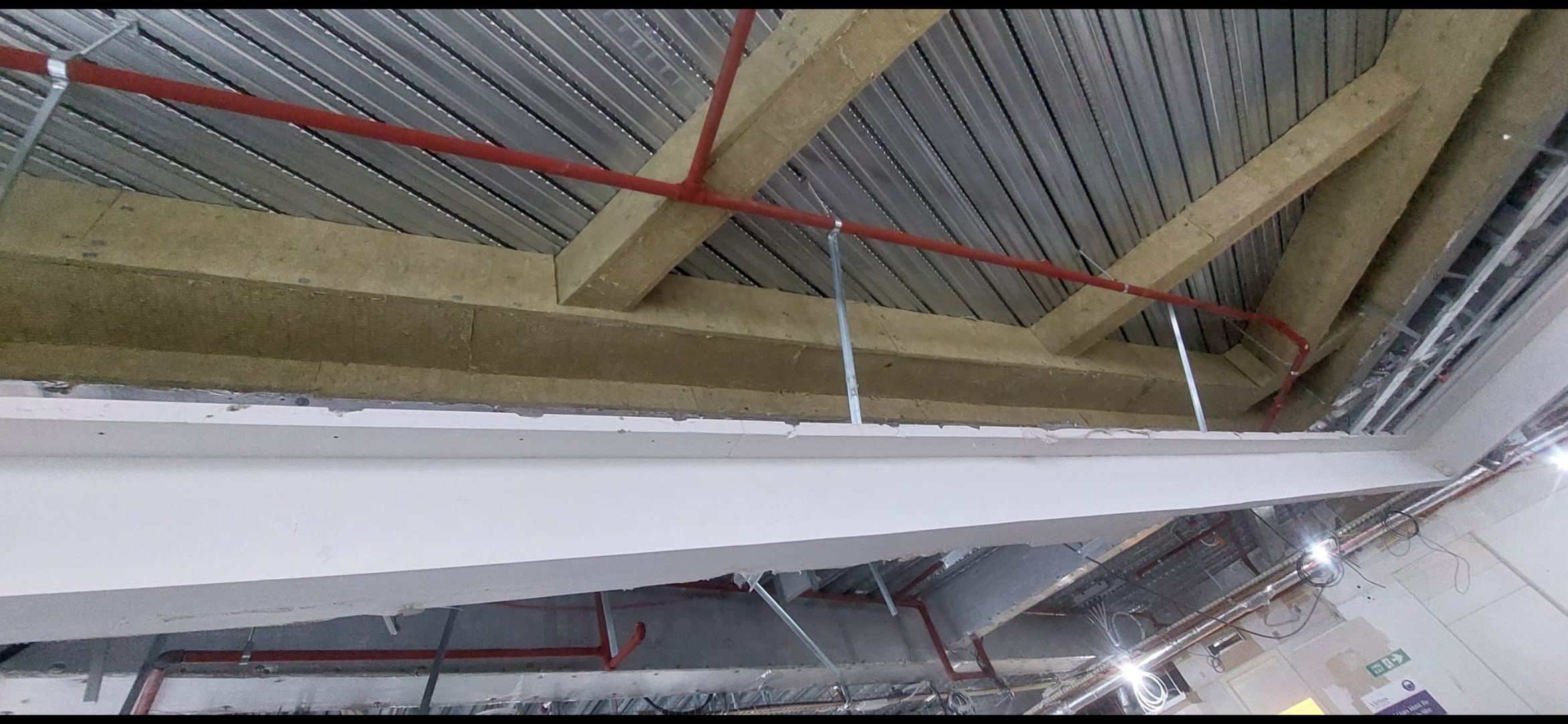 Exposed ceiling showing concrete beams, metal decking, and red fire sprinkler pipes during construction.