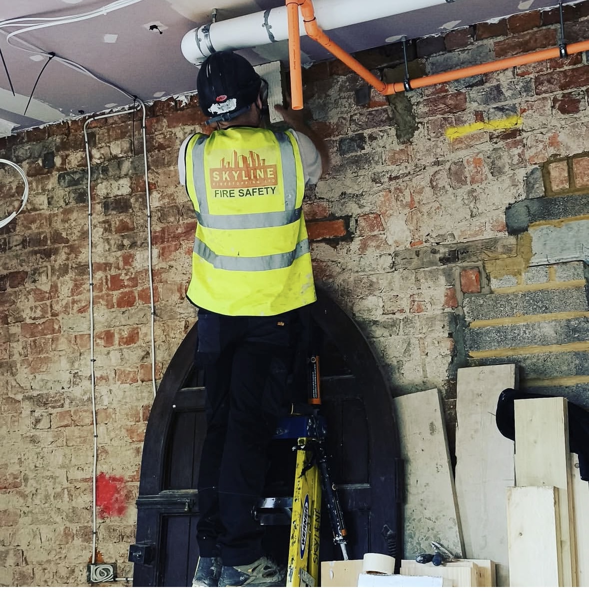 Worker in high-visibility vest working on pipes near ceiling in brick building.