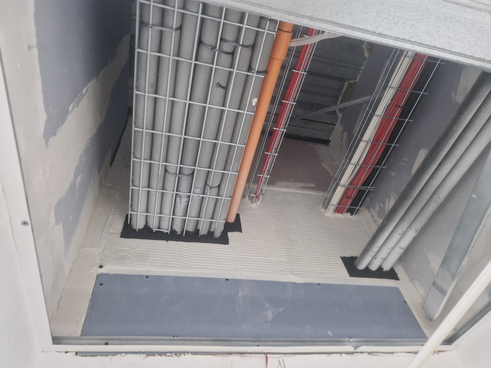 Service shaft with insulated pipes and cable trays passing through a floor penetration.